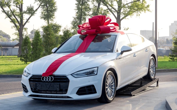 Luxury white Audi sedan with a giant red gift bow displayed outdoors — brand new premium car gift presentation.